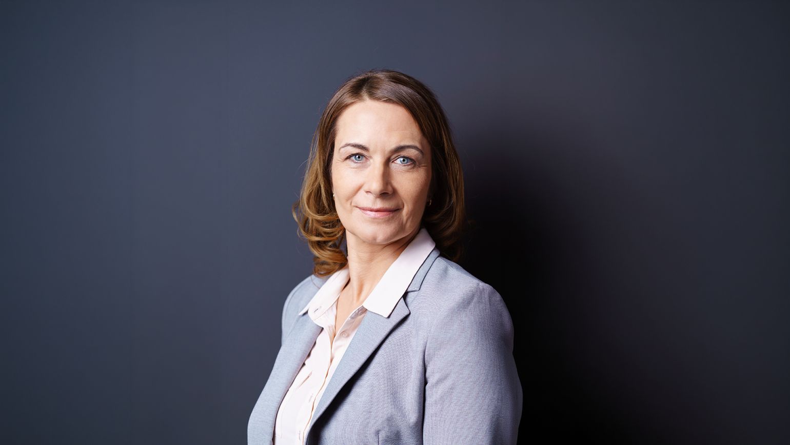 Studioaufnahme einer Business-F&uuml;hrungskraft vor dunkelgrauem Hintergrund, in hellgrauem Blazer und wei&szlig;er Bluse, mit direktem Blick in die Kamera. Das klare, sachliche Portr&auml;t vermittelt Vertrauen, Kompetenz und Seriosit&auml;t &ndash; ideal f&uuml;r B2B-Kommunikation in regulierten Branchen, Qualit&auml;tssicherung und Compliance.