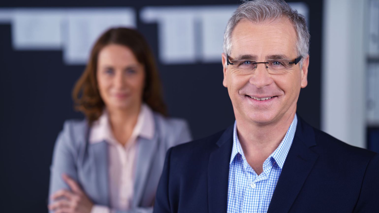 Portr&auml;t zweier Fachkr&auml;fte in Businesskleidung in einer modernen B&uuml;roumgebung: Ein l&auml;chelnder Manager im Vordergrund, eine Kollegin mit verschr&auml;nkten Armen unscharf im Hintergrund. Ideal zur Darstellung von Leadership, Auditbereitschaft, Compliance und verl&auml;sslicher Qualit&auml;tssicherung.