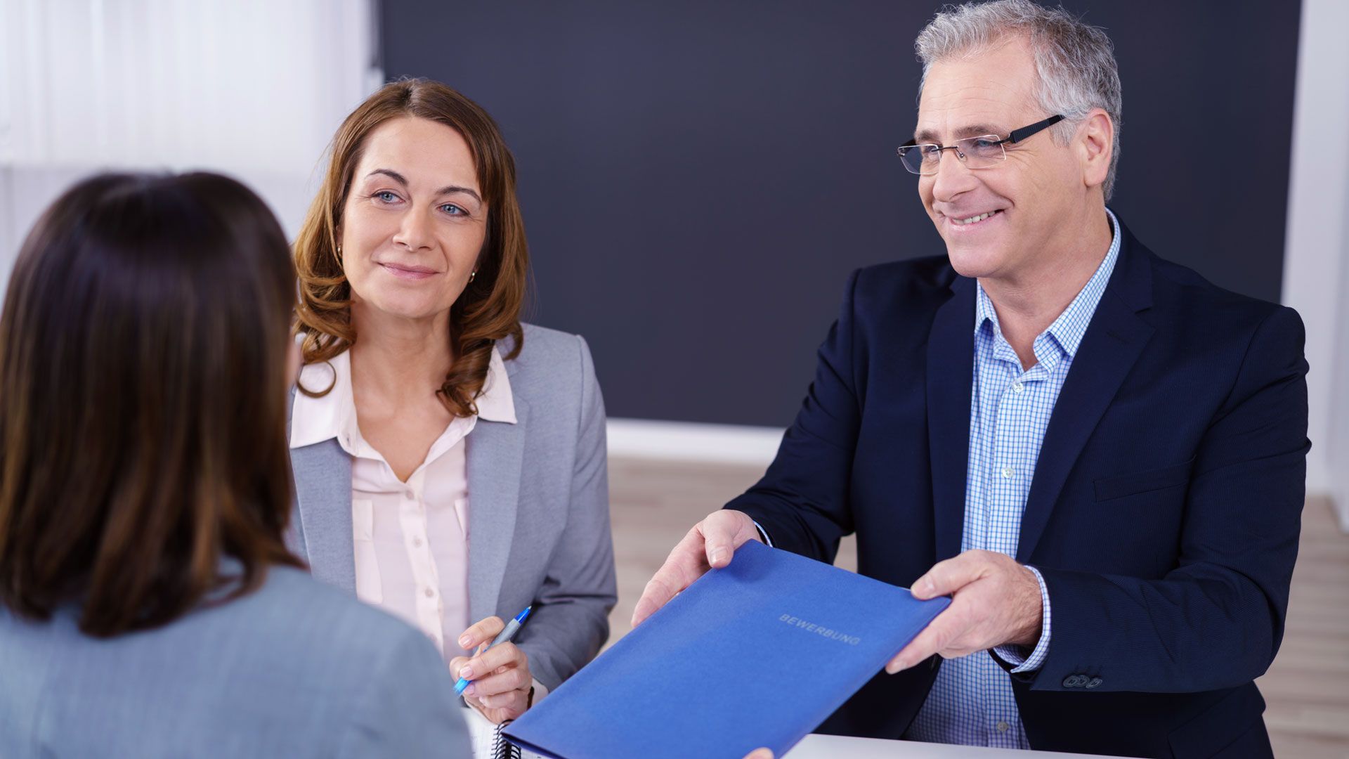 Zwei Fachpersonen &uuml;berreichen in einem Meeting einen blauen Dokumentenordner an eine Kundin im Vordergrund. Die Szene vermittelt strukturierte Zusammenarbeit, Verl&auml;sslichkeit und Compliance&mdash;ideal zur Illustration unabh&auml;ngiger Pr&uuml;fung, Auditvorbereitung und Zertifizierungsprozesse in regulierten, sicherheitskritischen Branchen.